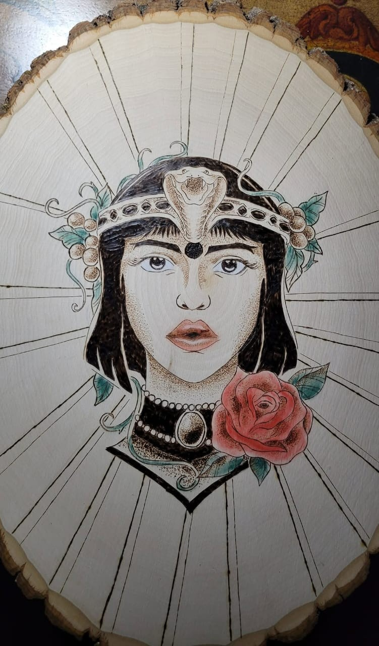 Pyrography wood-burn artwork on a circular wood slice of a woman's face with a cobra crown, roses, and teal floral details